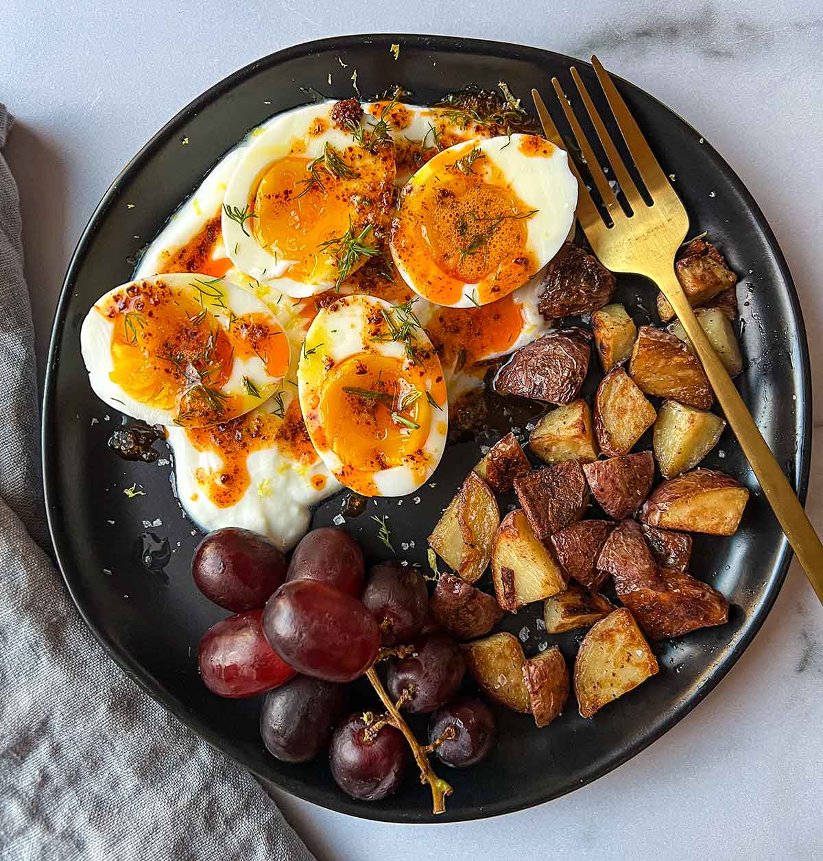 jammy eggs in yogurt roasted potatoes and red grapes on black plate with grey napkin and gold fork