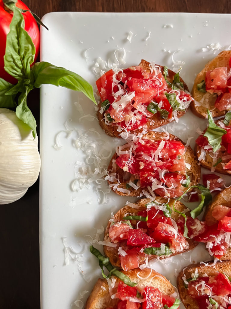 Easy Bruschetta with fresh red tomatoes, green basil on white serving platter with whole garlic