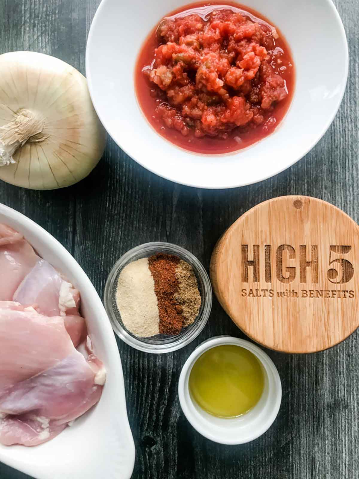 overhead shot of ingredients including whole onion, salsa, raw chicken, high 5 salts, spices and olive oil on a dark background