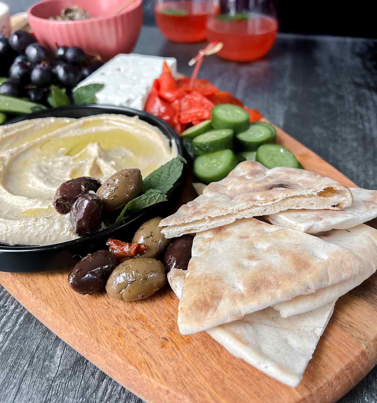 close up view of pita bread on wood cutting board with hummus, olives, cucumber, roasted red pepper and drink in background