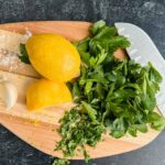 ingredients on oval cutting board with sharp knife and lemons, parsley and garlic on black counter