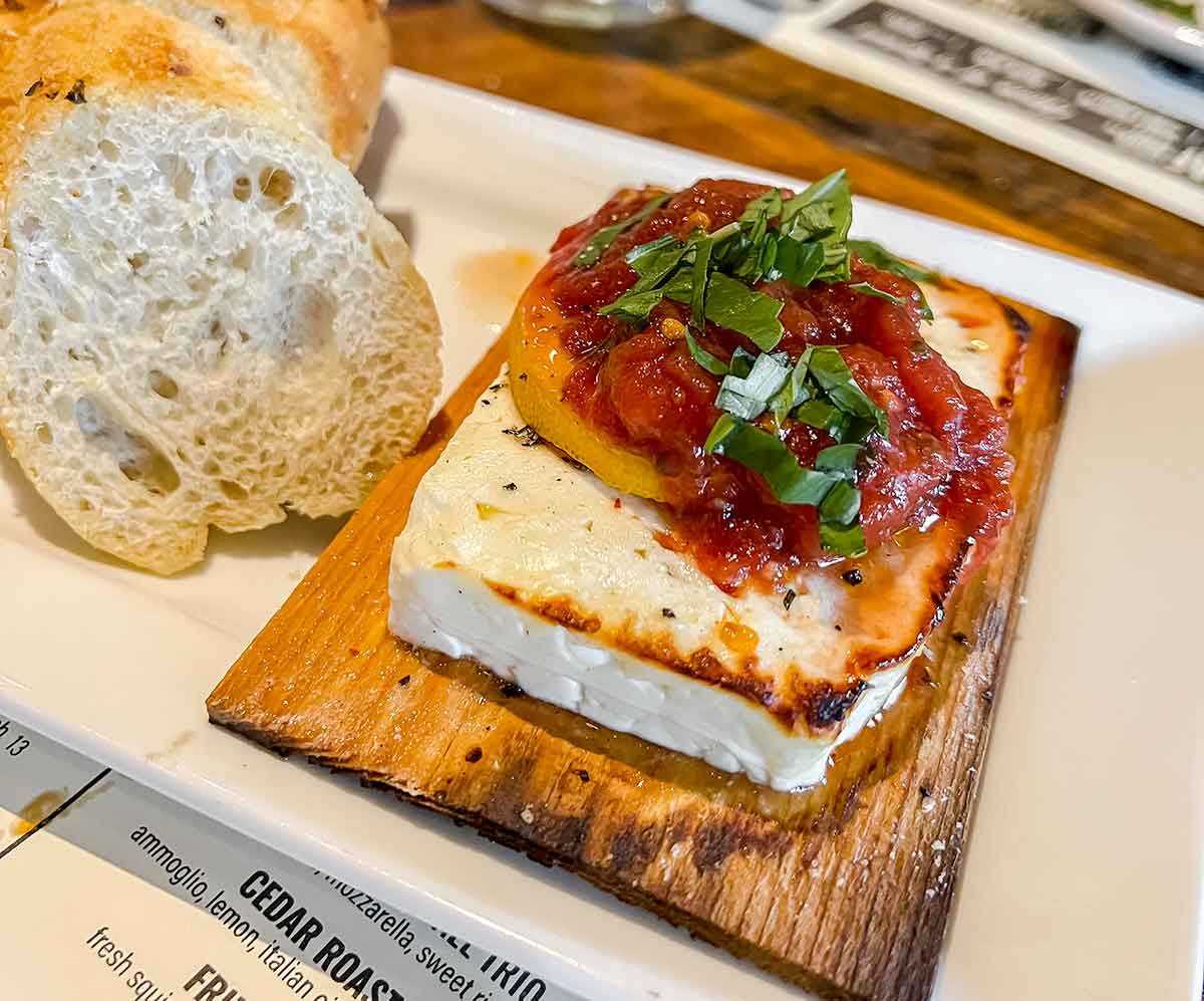 baked block of cheese on cedar plank topped with lemon and tomato sauce with sliced bread
