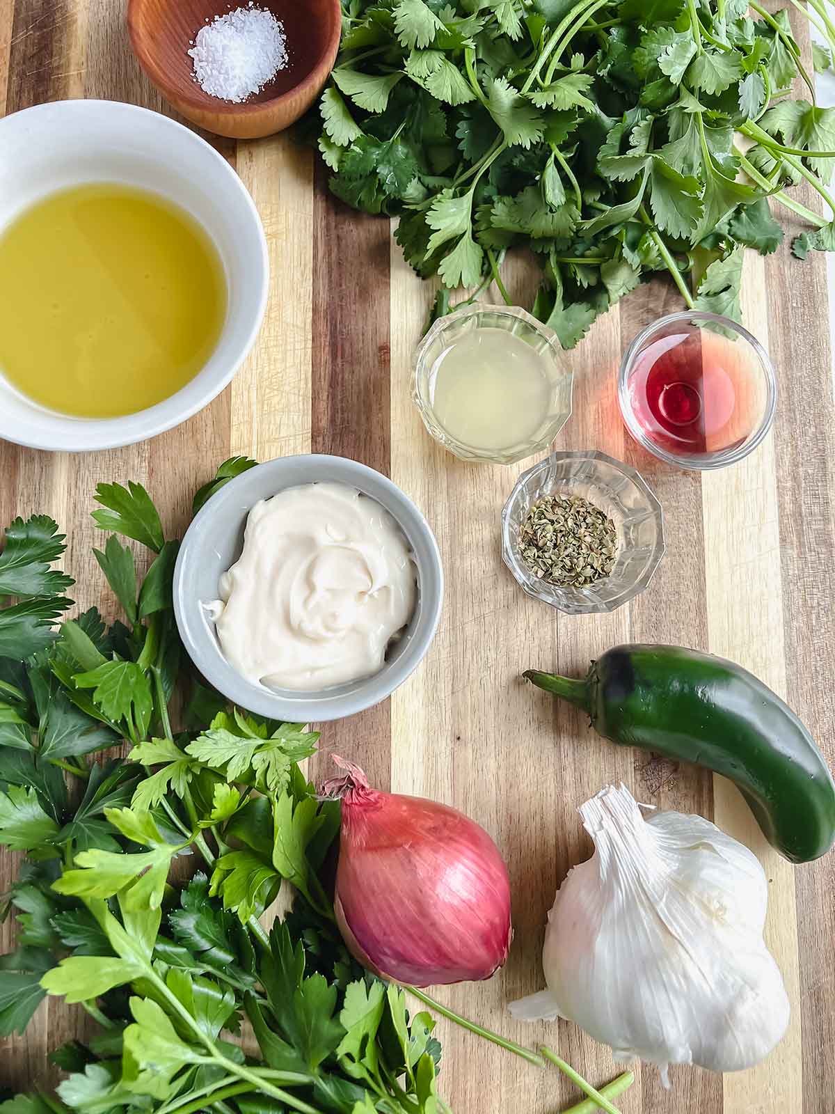 chimichurri aioli ingredients including garlic, parsley, cilantro, olive oil, kosher salt on wood cutting board