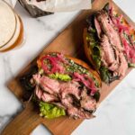 flank steak sandwich on wooden cutting board with beer and sweet potatoes
