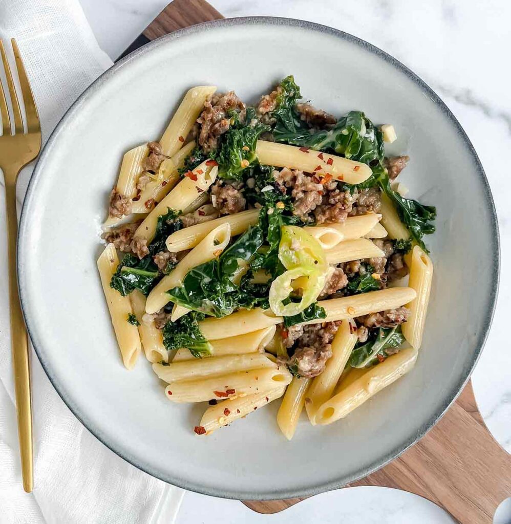 bowl of penne pasta with kale on wood cutting board and gold fork