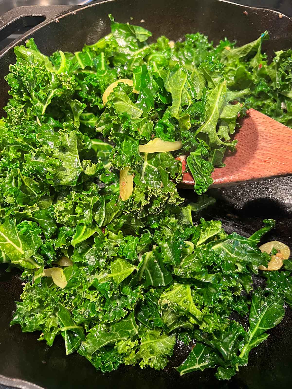 shredded kale and hungarian peppers cooking in cast iron with wooden spoon stirring